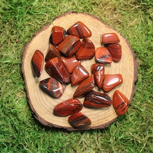 May include: A collection of red and black striped tumbled stones on a wooden slice. The stones are smooth and polished, with a variety of shapes and sizes.
