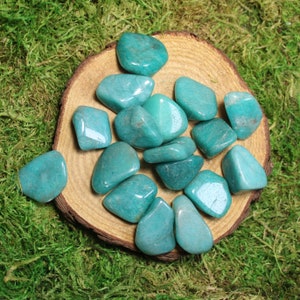 May include: A collection of smooth, teal green amazonite tumbled stones on a wooden surface. The stones are various sizes and shapes, and have a natural, polished finish.