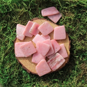 May include: A collection of pink opal stones, some with a striated pattern, arranged on a wooden surface with a green moss background.