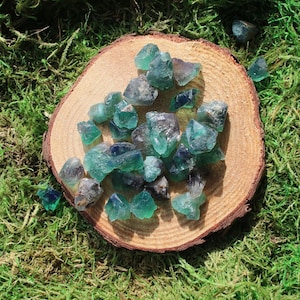May include: A collection of green and blue fluorite crystals on a wooden slice. The crystals are various sizes and shapes, with some having a more opaque appearance than others.