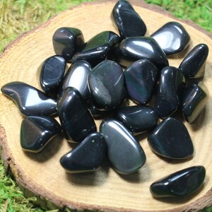 May include: A collection of black obsidian tumbled stones with iridescent rainbow flashes. The stones are smooth and polished, and they are arranged on a wooden surface.