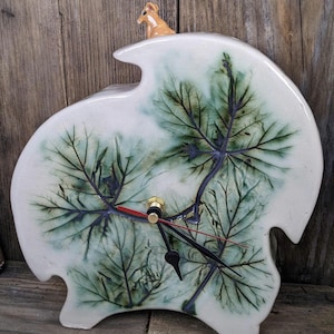 May include: A ceramic wall clock with a leaf design in shades of green and blue. The clock has black hands, a gold-coloured centre, and a small brown animal figurine on top. The clock is approximately 25 cm wide.
