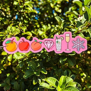 May include: A pink sticker featuring illustrations of an orange, apple, peach, diamond, cocktail, champagne flute, and snowflake. The word "TALLMADE" is printed on the cocktail illustration.