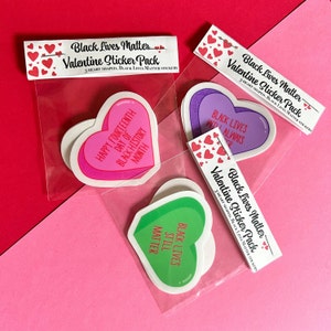 May include: A pack of three heart-shaped stickers with Black Lives Matter messages. The stickers are in a clear plastic bag with a header that reads "Black Lives Matter Valentine Sticker Pack". The stickers feature the messages "Black Lives Still Matter", "Black Lives With Always", and "Happy Fourteenth Day of Black History Month".