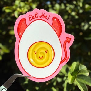 May include: A pink sticker featuring a devilish egg design. The egg has red devil horns and a tail, with the text "Eat Me!" in red. The egg yolk has a smiling face. The sticker is held by tweezers.