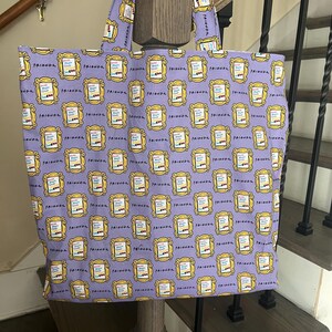 May include: A purple tote bag with a repeating pattern of yellow picture frames with the word "Friends" written inside. The frames are filled with colorful images of the characters from the TV show "Friends".