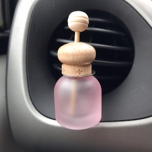 May include: A pink, frosted glass car air freshener with a wooden top and a small wooden ball on top. The air freshener is set against a car's air vent and dashboard.