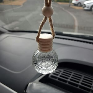 May include: A clear glass car air freshener bottle with a faceted geometric design. The bottle features a light-coloured wooden cap and is suspended by a tan jute rope with a matching round wooden bead, designed for hanging from a rearview mirror.