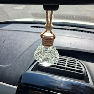 May include: A clear glass car air freshener bottle with a faceted, diamond-like texture. The bottle features a light-coloured wooden cap and a matching wooden bead, suspended by a thick, natural-toned jute twine cord for hanging from a mirror.