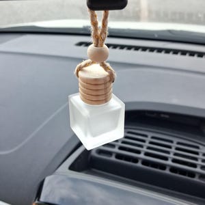 May include: A small, square, frosted glass bottle hanging from a rearview mirror by a braided jute cord. The bottle features a light-coloured, cylindrical wooden cap with horizontal grooves and a single round wooden bead above the cap.