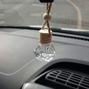 May include: A clear glass car air freshener bottle with a diamond-shaped base and a light-coloured wooden cap. The bottle hangs from a braided cord featuring a single round wooden bead, designed to be suspended from a vehicle rearview mirror.