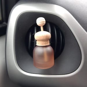 May include: A peach-coloured car air freshener with a wooden top and stick, placed in a car's air vent. The frosted glass bottle has a wooden stopper. The date and time, 15 October 2025 11:58, are visible in the bottom right corner.