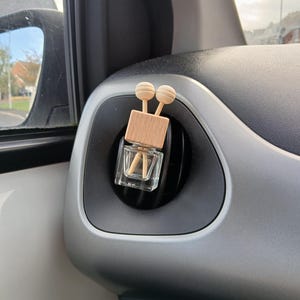 May include: A small, clear glass bottle with a wooden top and two wooden sticks inside. The bottle is hanging from a car's air vent.