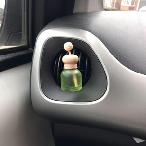 May include: A small, green glass car air freshener with a wooden cap and a wooden ball on top. The air freshener is placed in a car's air vent, surrounded by a black and silver dashboard.