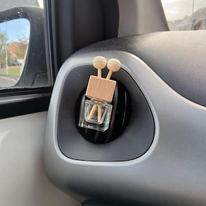 May include: A small, clear glass bottle with a wooden top and two wooden sticks inside. The bottle is hanging from a car's air vent.