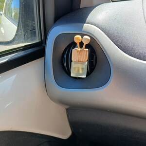 May include: A small, clear glass bottle with a wooden cap and two wooden sticks attached to the top. The bottle is hanging from a car's air vent.