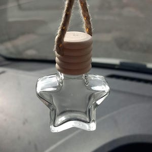 May include: A clear glass bottle shaped like a five-pointed star with a light-coloured wooden cap. The bottle is suspended by a thick, braided jute rope that is looped through the top of the wooden cap, allowing it to hang vertically.
