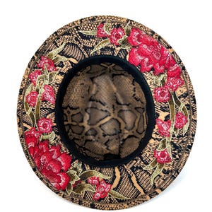 May include: A stylish hat with a snakeskin pattern and embroidered red flowers. The hat features a black brim and a snakeskin patterned interior. The floral embroidery includes red flowers and green leaves, adding a touch of elegance to the design.
