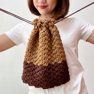May include: A brown and tan crocheted drawstring bag with a flower-like pattern. The bag is made of two different colours of yarn, with the top half being tan and the bottom half being brown.