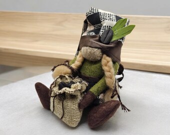 May include: A handmade gnome doll with a coffee theme. The gnome wears a green shirt, brown boots, and a black and white checkered hat. A brown strap holds the hat in place. The gnome has blonde braids and is holding a burlap sack.