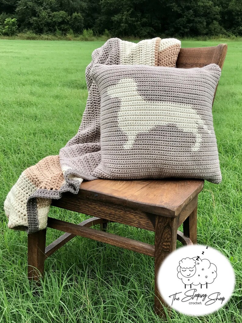May include: A crocheted pillow with a white dog silhouette on a light brown background. The pillow is sitting on a wooden chair with a crocheted blanket draped over the back. The chair is on a grassy lawn.