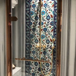 Pode incluir: Um box de chuveiro com uma porta de vidro e acess&oacute;rios dourados. O chuveiro &eacute; de design circular com acabamento dourado. O box de chuveiro &eacute; revestido com azulejos brancos com um padr&atilde;o floral azul e vermelho.
