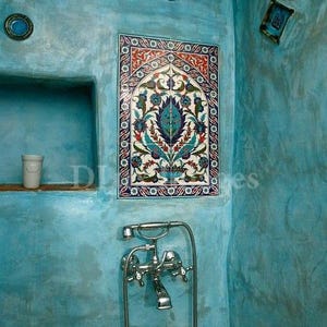 May include: A blue tiled bathroom with a decorative floral tile design. The tile is framed in red and white. A silver shower faucet is visible in the lower right corner.