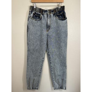 May include: High-waisted acid wash denim jeans featuring a gathered elastic waistband, a front button and zipper closure, two side pockets, and vertical zippered slits at the bottom of each leg hem, showcasing a light blue and white mottled pattern.