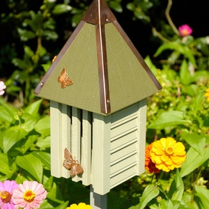 May include: A green and brown butterfly house with a copper roof. The house is mounted on a wooden post and surrounded by pink and yellow flowers.