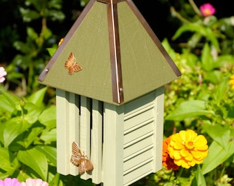 May include: A green and brown butterfly house with a copper roof. The house is mounted on a wooden post and surrounded by pink and yellow flowers.