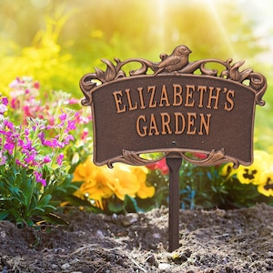 May include: A brown metal garden sign with an ornate design and the text "Elizabeth's Garden". The sign is attached to a metal stake and is placed in a garden bed with flowers.