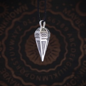 Puede incluir: Collar con colgante de plata con un dije en forma de pirámide con un diseño de ojo. El dije cuelga de un cordón negro.