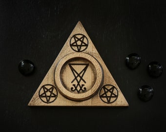Puede incluir: Un quemador de incienso triangular de madera con una depresión circular central y cuatro pentagramas tallados. El quemador está sobre una superficie negra con piedras negras alrededor.