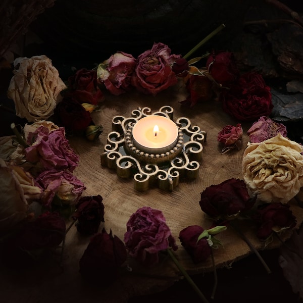 May include: A small, ornate, gold-coloured candle holder with a lit candle in the centre, surrounded by dried roses in shades of pink, red, and cream on a wooden surface.