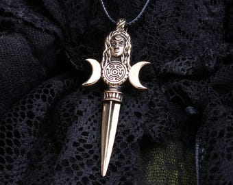 May include: A bronze-colored pendant necklace featuring a stylized female face, a circular maze design, and two crescent moons. The pendant has a dagger-like shape and hangs on a black cord, set against a dark, textured background.