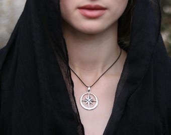 May include: A silver compass pendant necklace with a black cord. The pendant features a star design within a circular frame. The person in the image is wearing a black hooded garment.