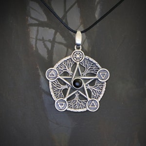 Może przedstawiać: Srebrny wisiorek z pięcioramienną gwiazdą z czarnym kamieniem w środku. Gwiazda jest otoczona kółkiem z wzorem drzewa życia i czterema symbolami żywiołów.