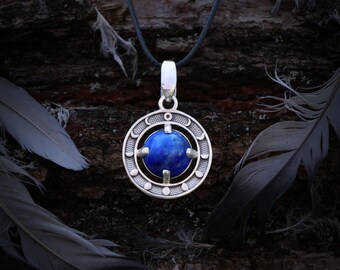 May include: A silver pendant with a round, deep blue stone at its center. The pendant has a circular design with crescent moon and dot details. It hangs from a dark gray cord, with feathers in the background.