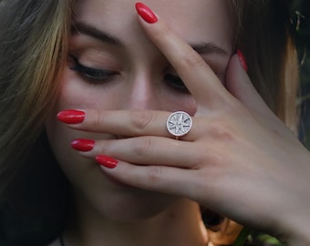 May include: A silver ring featuring a compass rose design. The ring has a circular face with a starburst pattern and a beaded border. The ring is worn on a finger with red nail polish.