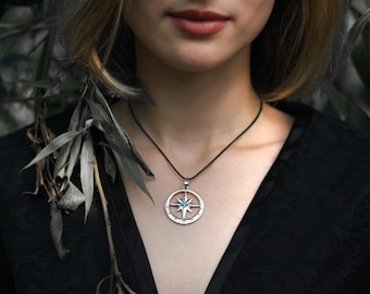 May include: A silver compass pendant with a blue gemstone center hangs from a black cord necklace. The pendant is circular with a star design inside. The person is wearing a black top.