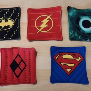 May include: Five square fabric coasters featuring superhero logos. Designs include Batman, The Flash, Green Lantern, Harley Quinn, and Superman. Each coaster has a different color scheme and logo.
