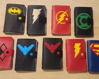 May include: A collection of nine rectangular leather-like journals, each featuring a unique superhero or comic book character emblem on the front cover. The journals are secured with a small snap-button strap and display various colors including black, red, and blue.