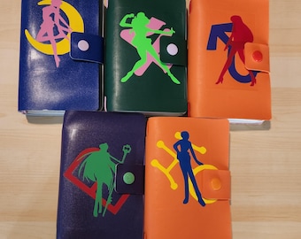 May include: Five rectangular notebooks with snap closures, each featuring a unique silhouette graphic. The covers are blue, dark green, orange, purple, and light orange, displaying colorful character designs including a crescent moon and symbols.
