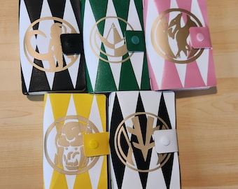 May include: Five rectangular wallets featuring a diamond pattern in black, green, pink, yellow, and white. Each wallet has a gold circular emblem with a unique animal or geometric design and a matching snap button closure on the right side.