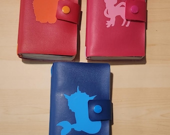 May include: Three rectangular notebooks with snap closures. The top left is red with an orange creature silhouette. The top right is magenta with a light pink creature silhouette. The bottom is blue with a light blue creature silhouette.