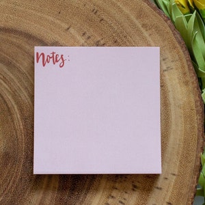 May include: A square, pale pink sticky note with the word "Notes" in reddish-brown script in the upper left corner. The note is on a wooden surface, with yellow and green floral accents in the background.