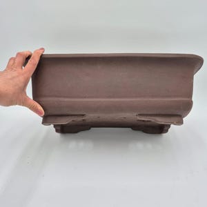 Peut inclure: Un pot à bonsaï rectangulaire marron avec un bord légèrement incurvé. Le pot a une surface texturée et est soutenu par quatre petits pieds. Le pot est conçu pour la culture des bonsaïs.