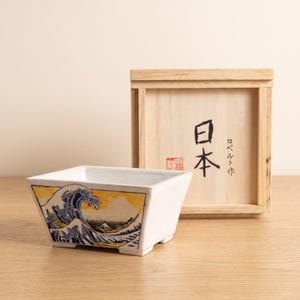 May include: A white ceramic rectangular bowl featuring a hand-painted blue and yellow wave design on the exterior. Behind the bowl sits a light-colored wooden box with the Japanese characters 日本 and ロベルト作 printed in black ink on the front.