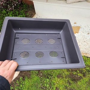 Peut inclure: Un bac à plantes rectangulaire gris foncé avec un insert perforé. Le bac est de forme carrée et est tenu par une main. L'insert comporte six ouvertures circulaires. Le bac est à l'extérieur, avec de l'herbe verte et des plantes en arrière-plan.