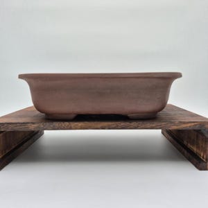 Może przedstawiać: Brązowa, prostokątna doniczka do bonsai z zaokrąglonymi krawędziami, spoczywająca na ciemnej drewnianej podstawce. Doniczka ma lekko zakrzywiony kształt i stoi na trzech małych nóżkach. Podstawka wykonana jest z ciemnego, bejcowanego drewna.